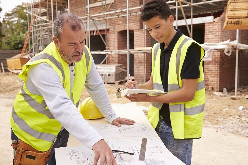 Builder On Building Site Discussing Work With Apprentice cm