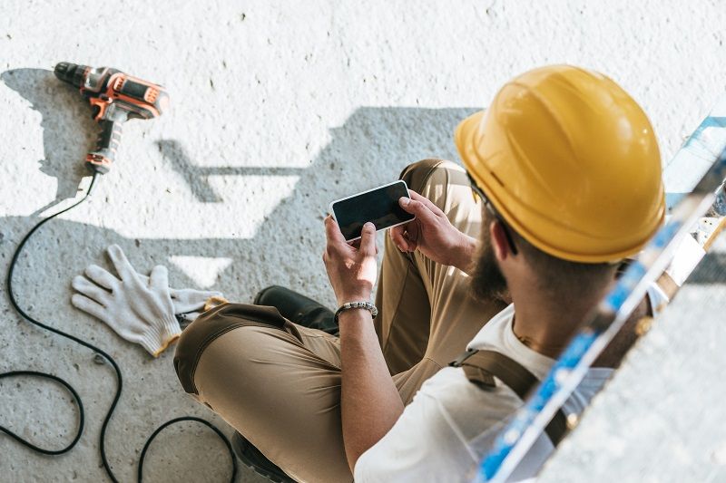overhead-view-of-builder-in-protective-helmet-using-smartphone-with-blank-screen-at-construction-site-cm