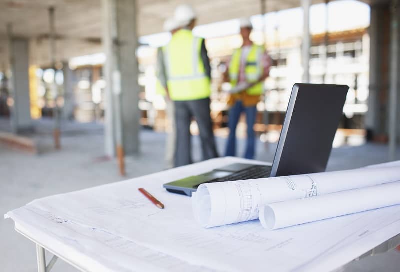 Construction workers behind blueprints and laptop on construction site-cm