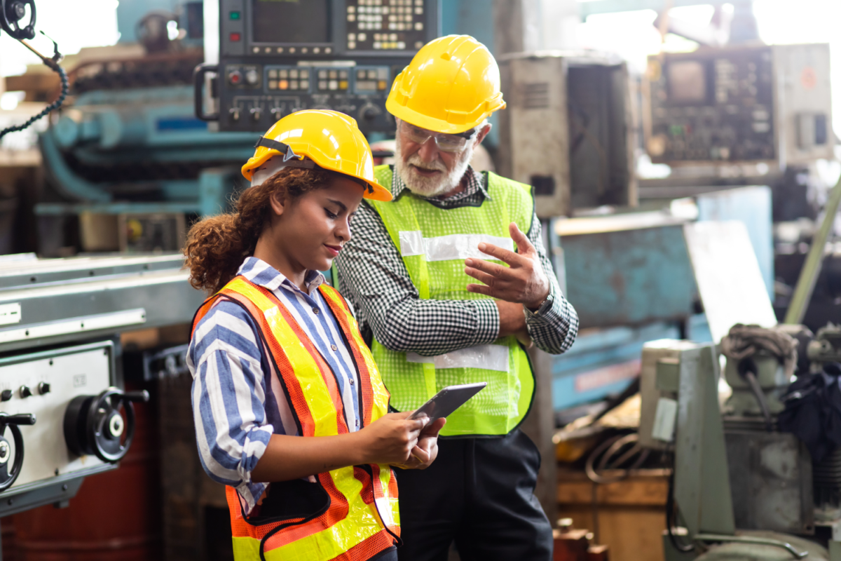 Two manufacturing workers in safety gear reviewing information on a tablet in a factory setting, illustrating how staffing agencies support industrial hiring by providing skilled, ready-to-work employees.