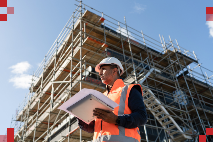 Construction worker in a white hard hat and orange safety vest holding a clipboard, standing in front of a multi-story scaffolding structure under a clear blue sky.